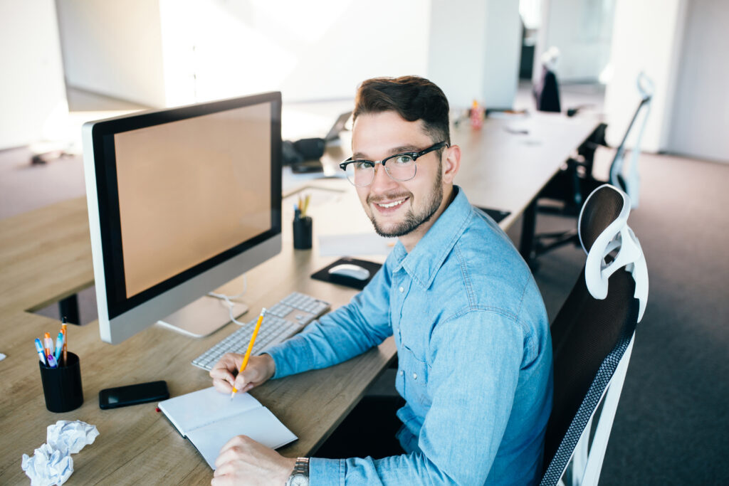 Young man in glassess is working at his workplace in office. He wears blue shirt. He is writing in notebook and smiling to the camera Young man in glassess is working at his workplace in office. He wears blue shirt. He is writing in notebook and smiling to the camera